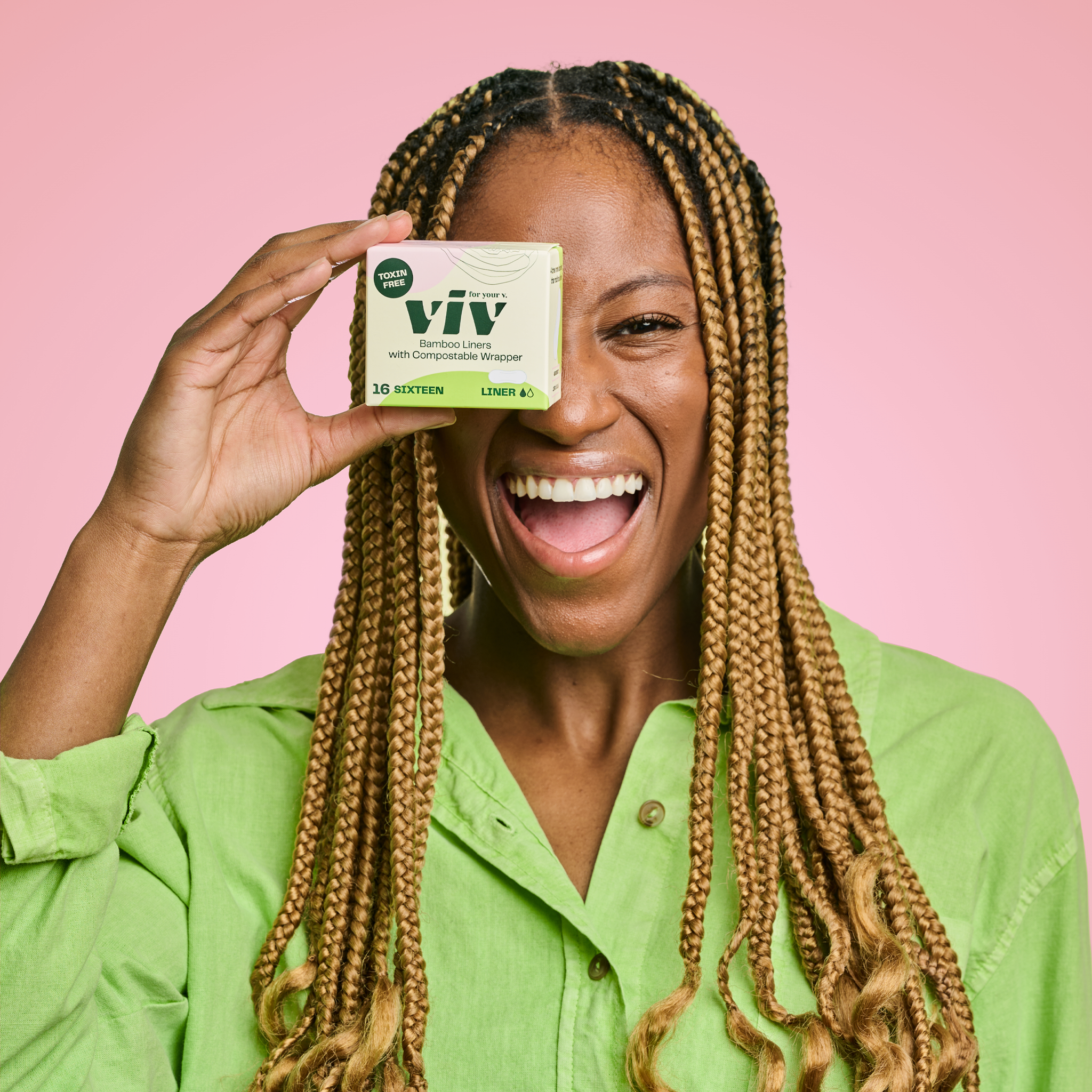 Gen-Z woman in a green shirt holding a box of panty liners in front of her eye, she is smiling and looking content.