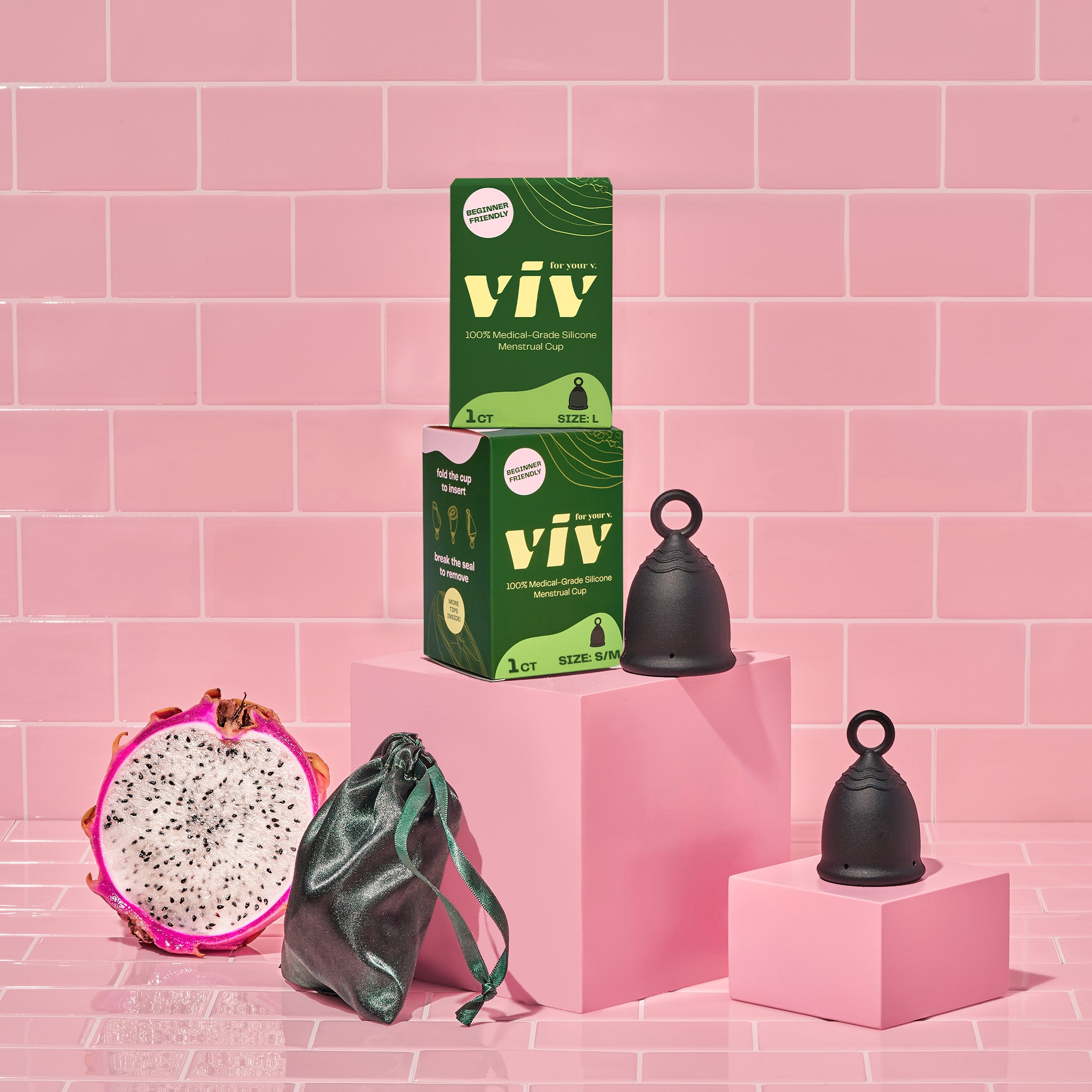 Two viv menstrual cups sit on pink boxes in a pink tiled room. Two viv period cup boxes are stacked to the left. A menstrual cup pouch sits in the front with a dragonfruit next to it.
