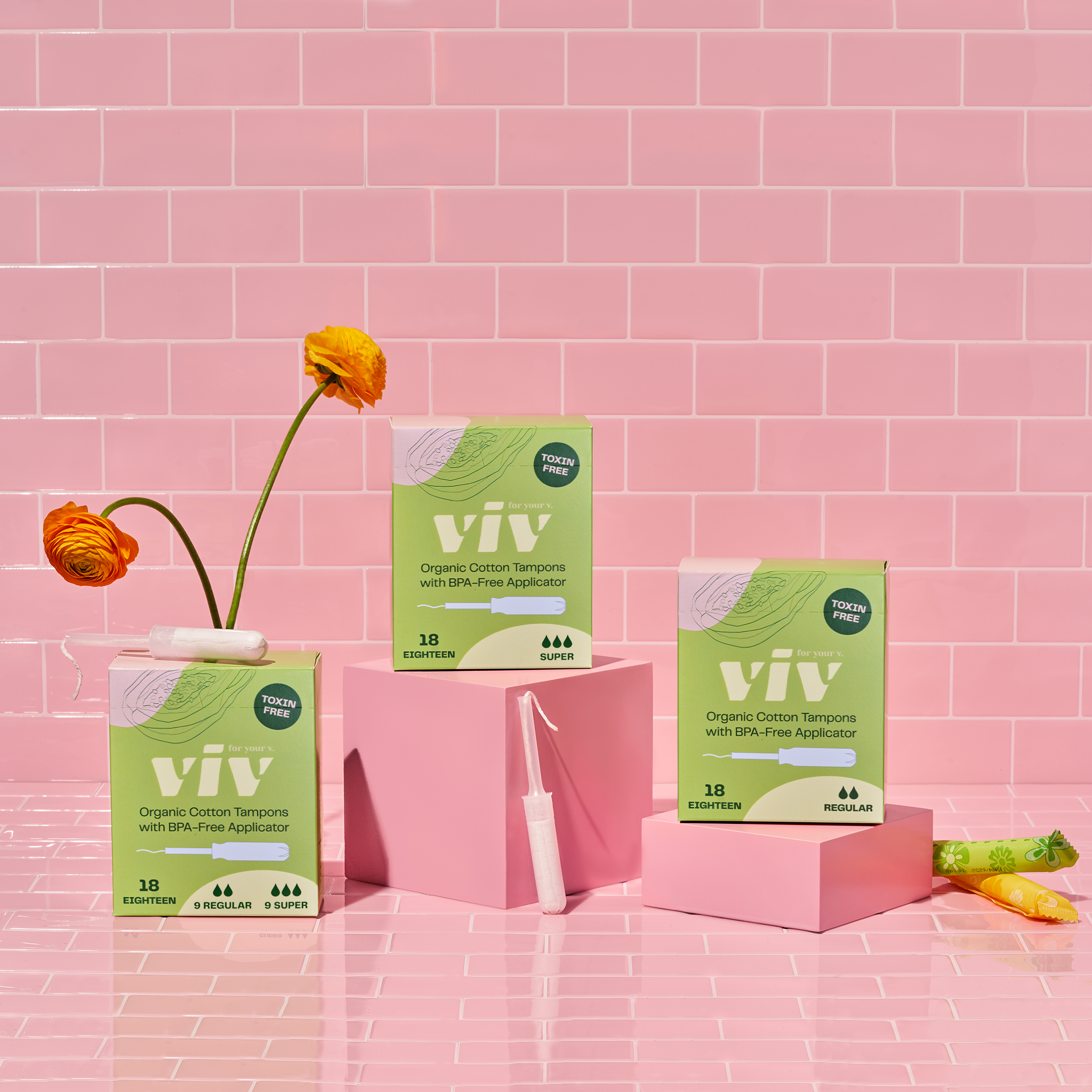 Three boxes of viv organic cotton tampons with BPA-Free applicator sitting in a pink tiled room with flowers in the background. A sustainable tampon stands in front.