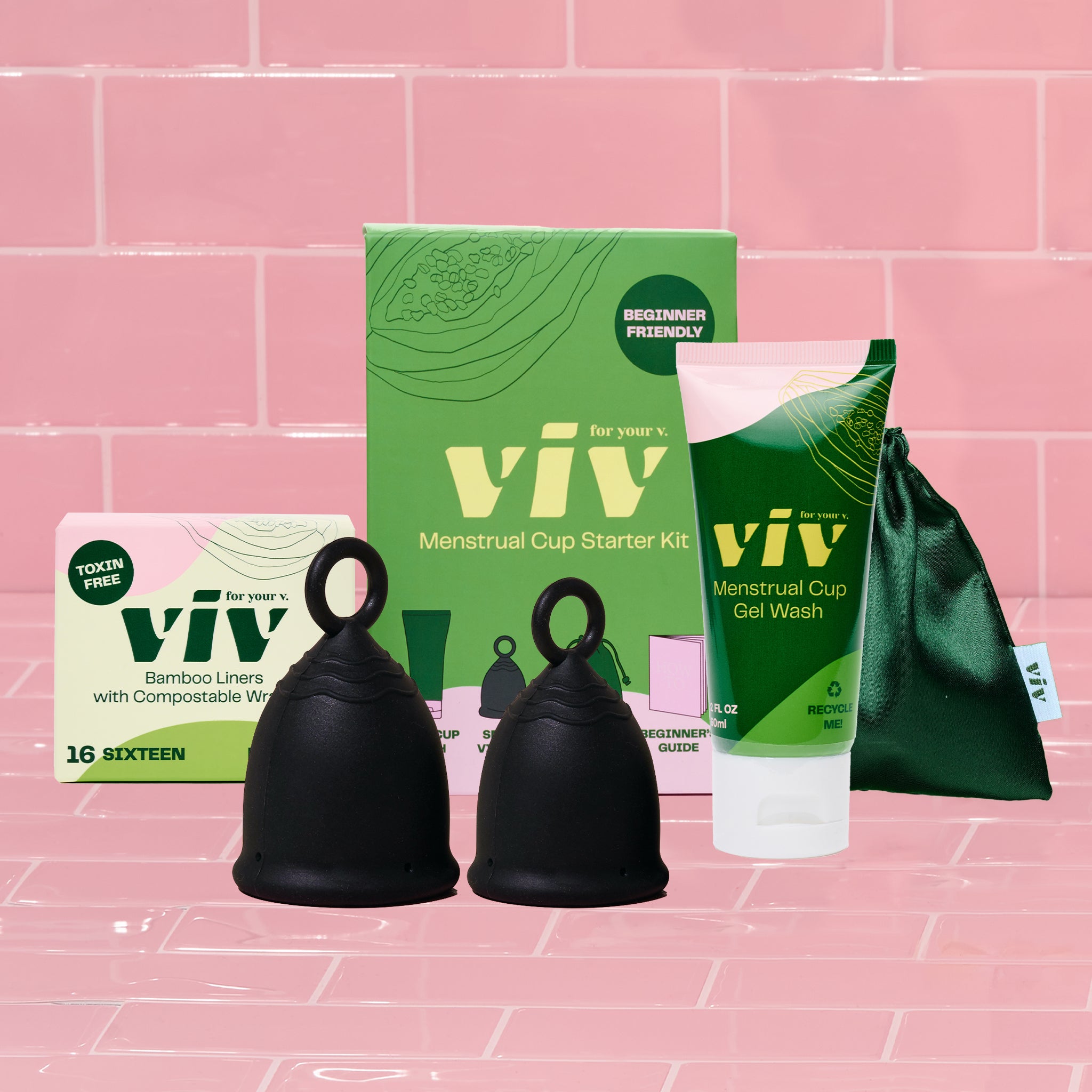 Beginner Friendly Viv Menstrual Cup Perfect Fit Kit. Two black viv menstrual cups sit in front in a pink tiled room. Behind the cups are a box of sustainable bamboo liners, the menstrual cup starter kit box, an all natural menstrual cup gel wash, and the emerald green viv period cup pouch.