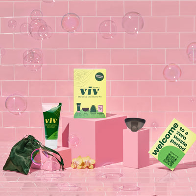 The Viv Menstrual Disc Starter Kit sits on a pink tile background, with a Viv Menstrual Disc Cleanser, the Small Viv Disc, a Menstrual Disc Beginner's guide reading "Welcome to a zero waste period," and a green silk storage pouch to store your Viv Menstrual Disc