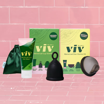 The Viv Menstrual Cup starter kit and Viv Menstrual Disc Starter Kit sit on a pink tile background, with the Viv Disc and Viv Cup leaning against their boxes, alongside the Viv Menstrual Cup and Disc Gel Wash and a green silk storage pouch for your cup and disc